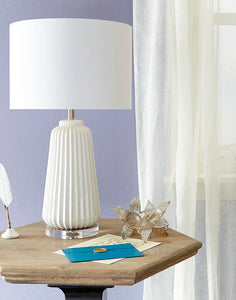 Experience the texture and refinement of sleek, smooth ridges with the Delphine Lamp. This ceramic table lamp is perfect for adding charm to a side table or a nightstand. Its crystal base and white finish over ceramic construction provide an engaging design. Additionally, the lamp features a white drum shade with a white lining to continue a monochromatic theme, achieving a clean and composed look that perfectly complements modern living.