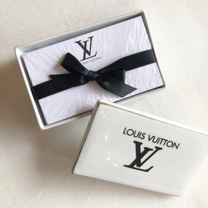 Impress your connections with this ceramic notecard box adorned with a chic fashion insignia. Accompanied by a set of matching notecards, this set is the epitome of tasteful luxury and elegance. It's the perfect way to leave a lasting impression with your personal touch.